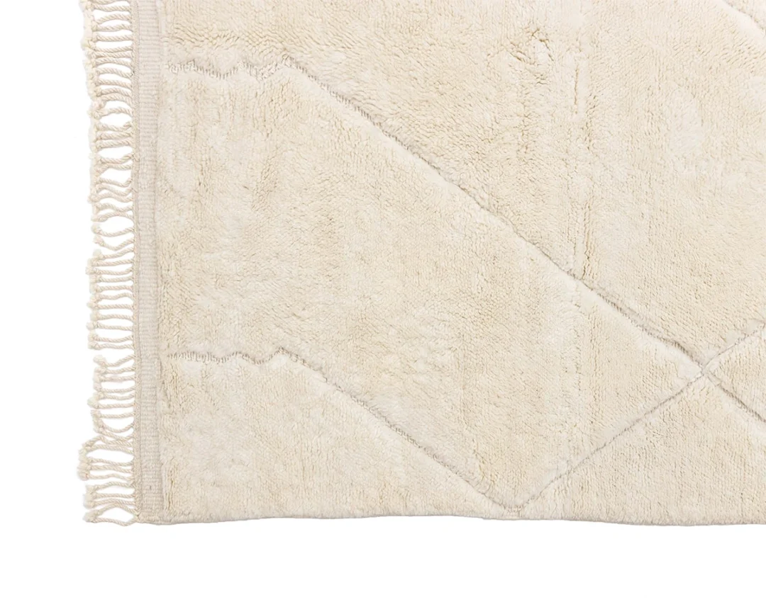 Close-up of the corner of a beige rug with a geometric pattern and fringe along the edge on a white background.