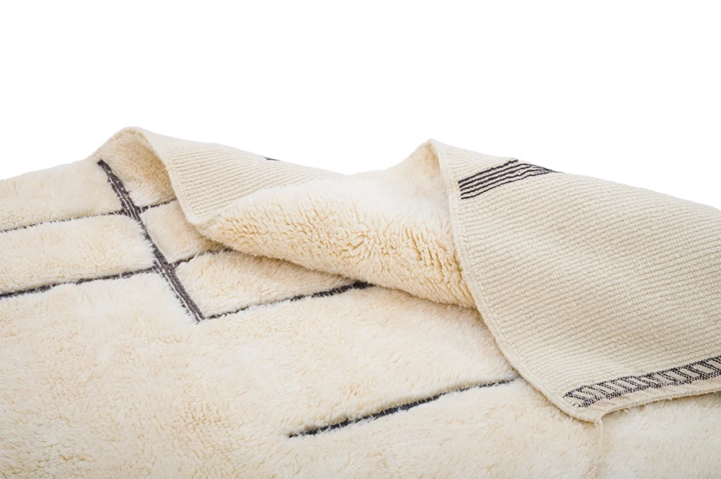 A close-up of a cream-colored, textured rug with black line patterns, partially folded to show both the front and back sides.