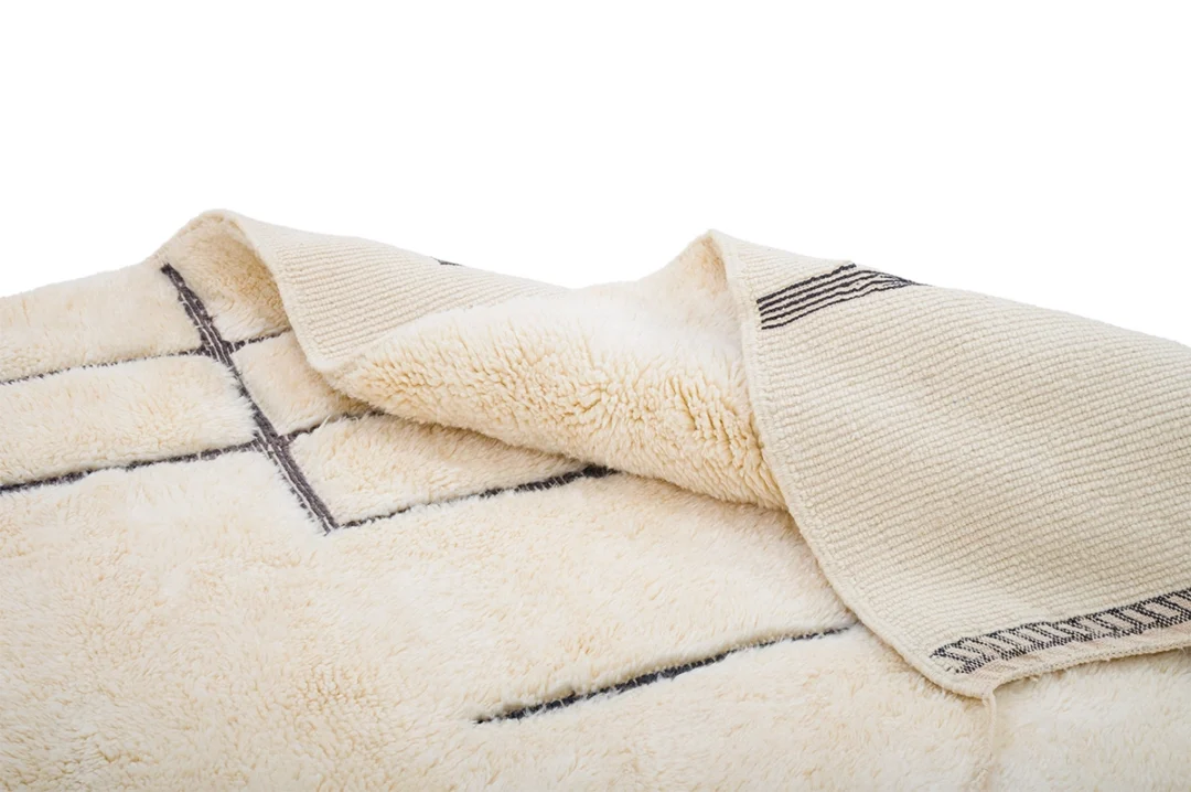A close-up of a cream-colored, textured rug with black line patterns, partially folded to show both the front and back sides.