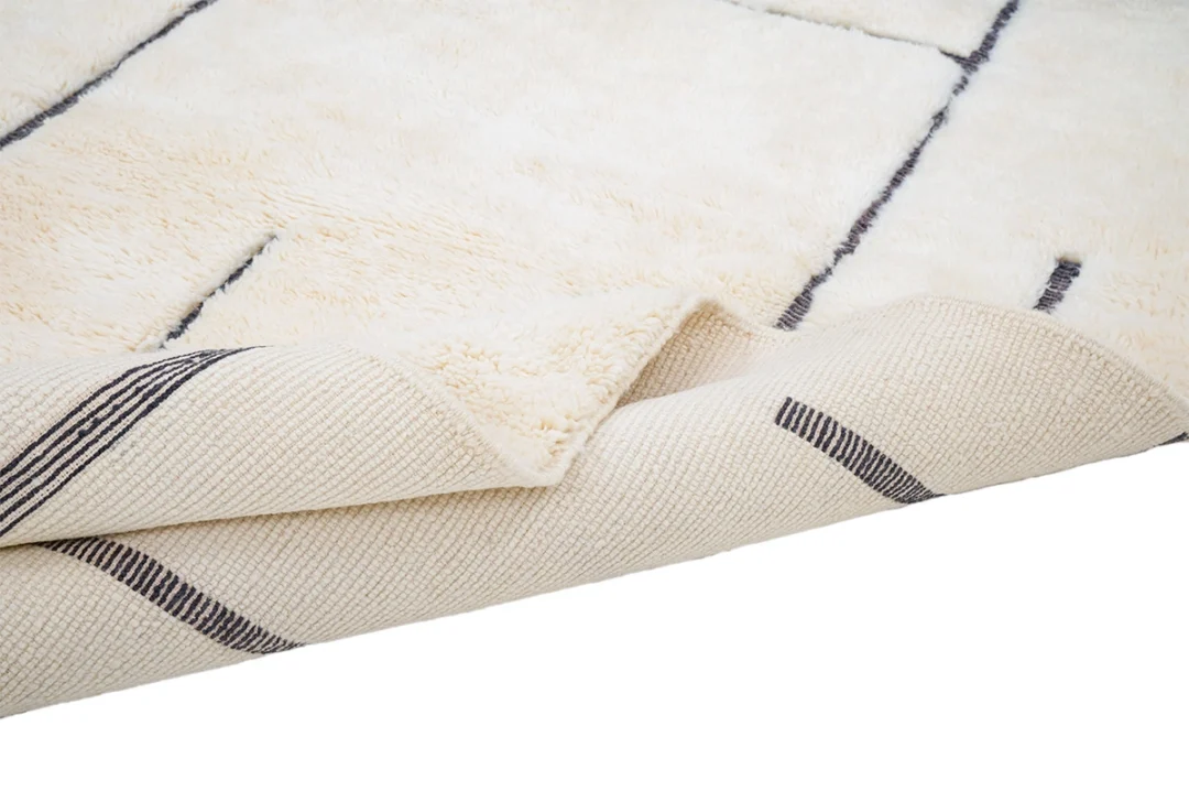 A close-up of a cream-colored area rug with thin black lines, showing the textured front and woven underside with part of the corner folded over.