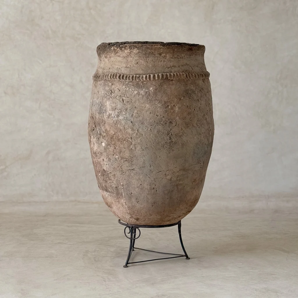 A large, weathered clay pot with a decorative band near the rim sits on a simple metal stand against a neutral background.
