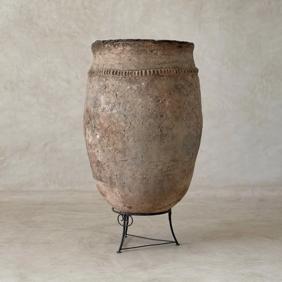 A large, weathered clay pot with a decorative band near the rim sits on a simple metal stand against a neutral background.
