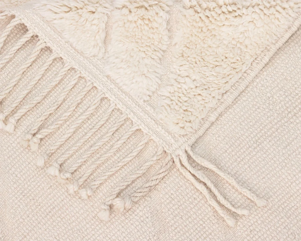 Close-up of a cream-colored woven textile with a textured, fluffy section and a row of twisted fringe tassels along the edge.