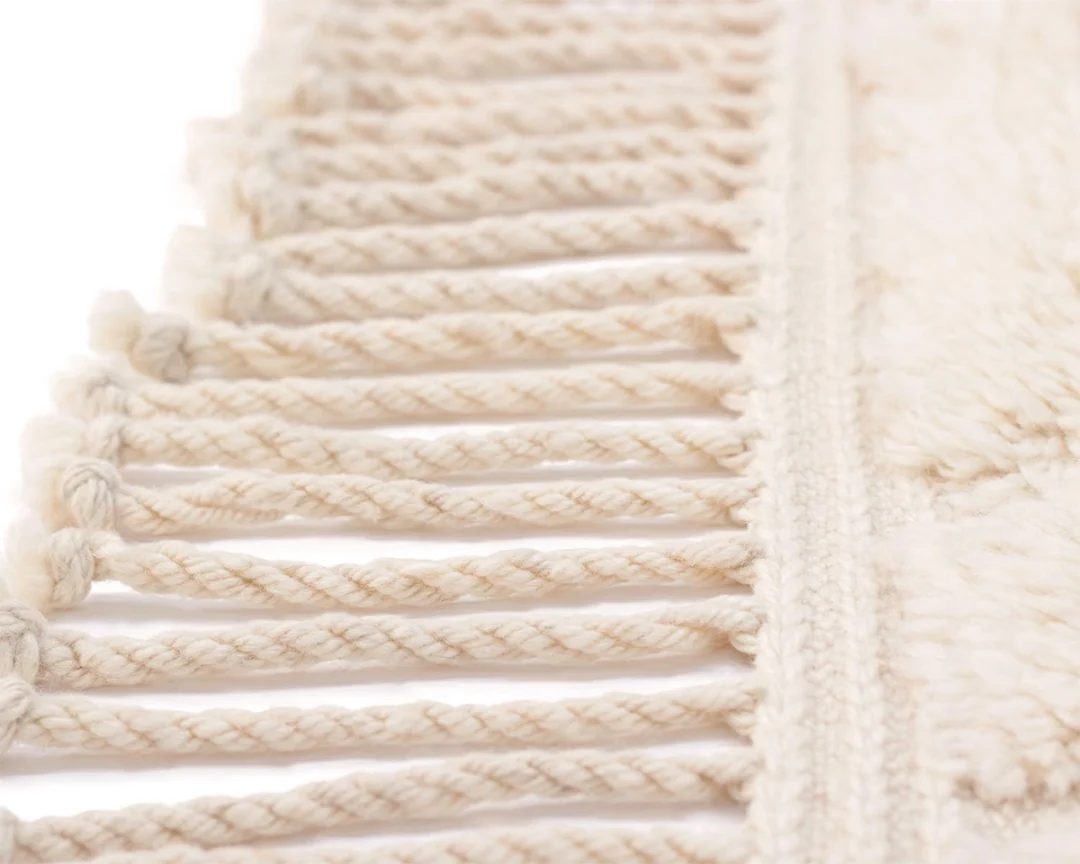 Close-up of cream-colored twisted fringes on the edge of a knotted rug, set against a white background.