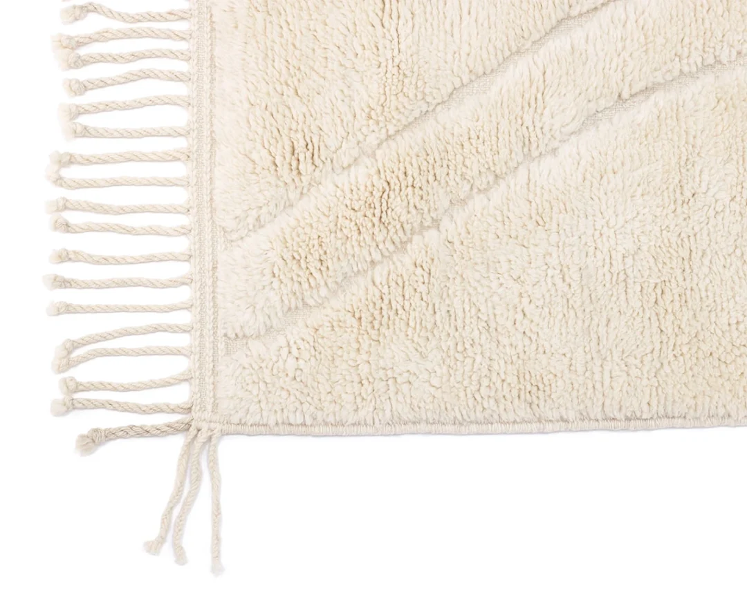 Close-up of a cream-colored, textured rug corner with diagonal lines and fringed edges.