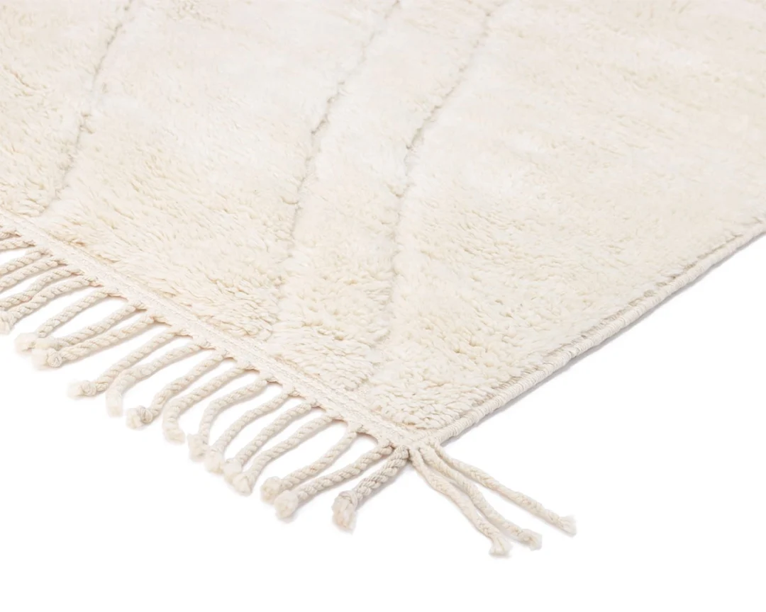 Close-up of a cream-colored, textured rug with knotted fringe tassels along the edge, shown on a white background.