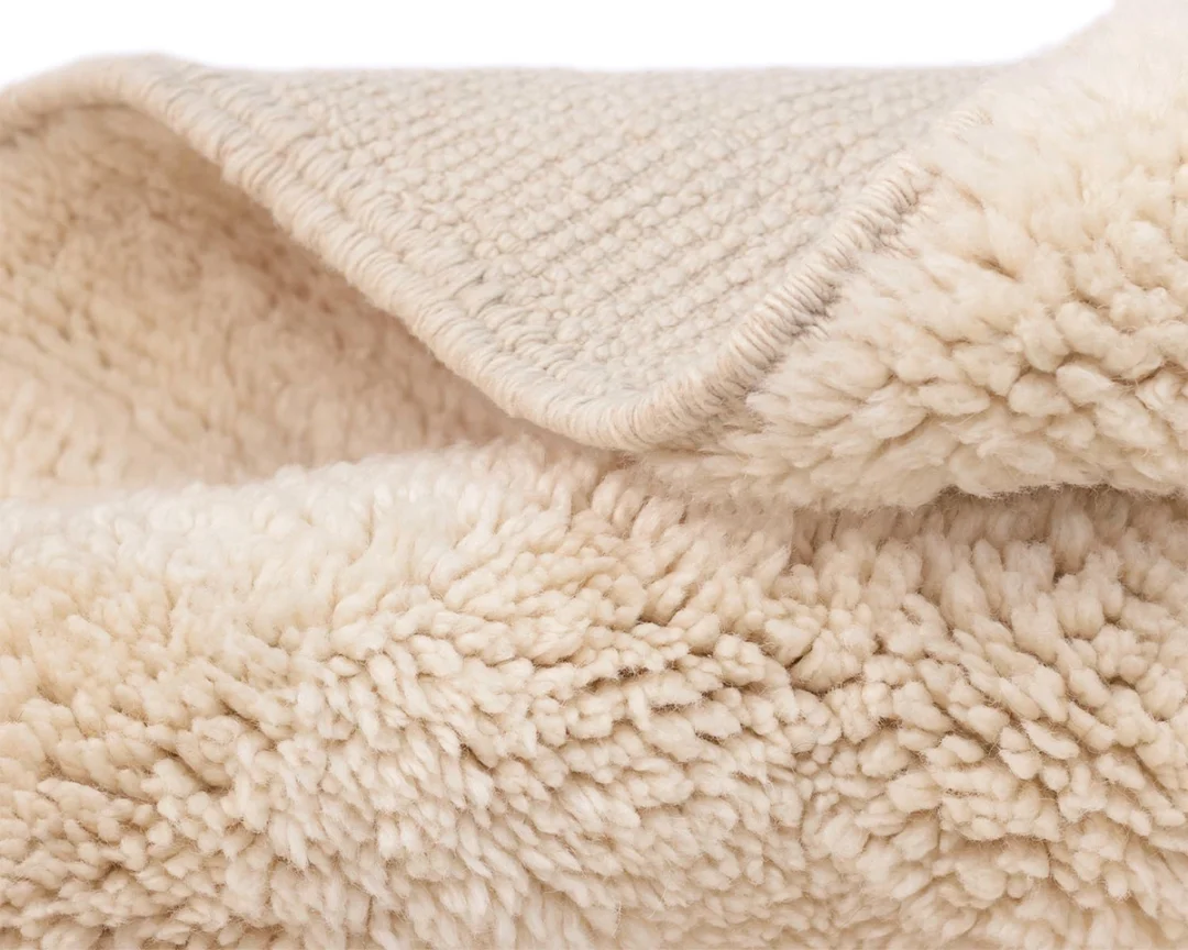 Close-up of a folded, beige, fluffy fleece or sherpa rug showing its soft, textured surface and stitched edge.