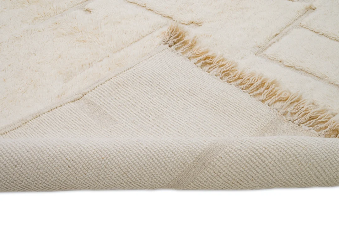 A close-up view of a cream-colored textured rug with a corner flipped over, revealing the woven underside and frayed edge.