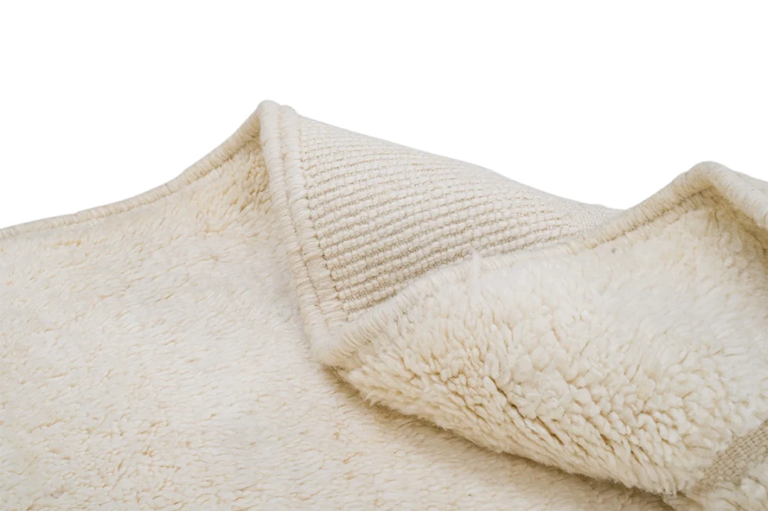 A close-up of a cream-colored, textured rug with a soft, fluffy surface and a visible woven underside, partially folded over on a white background.