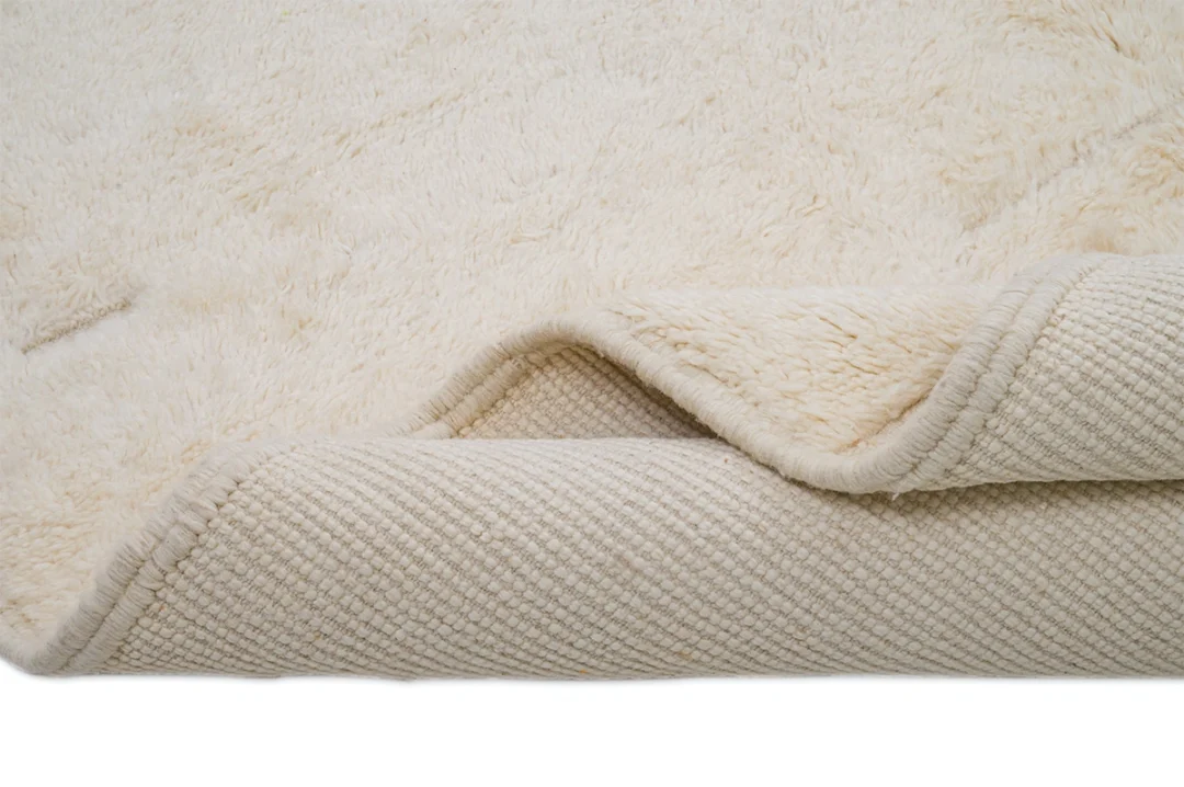 A close-up of a cream-colored, textured rug with a soft, fluffy surface and the corner folded over to reveal a woven underside.