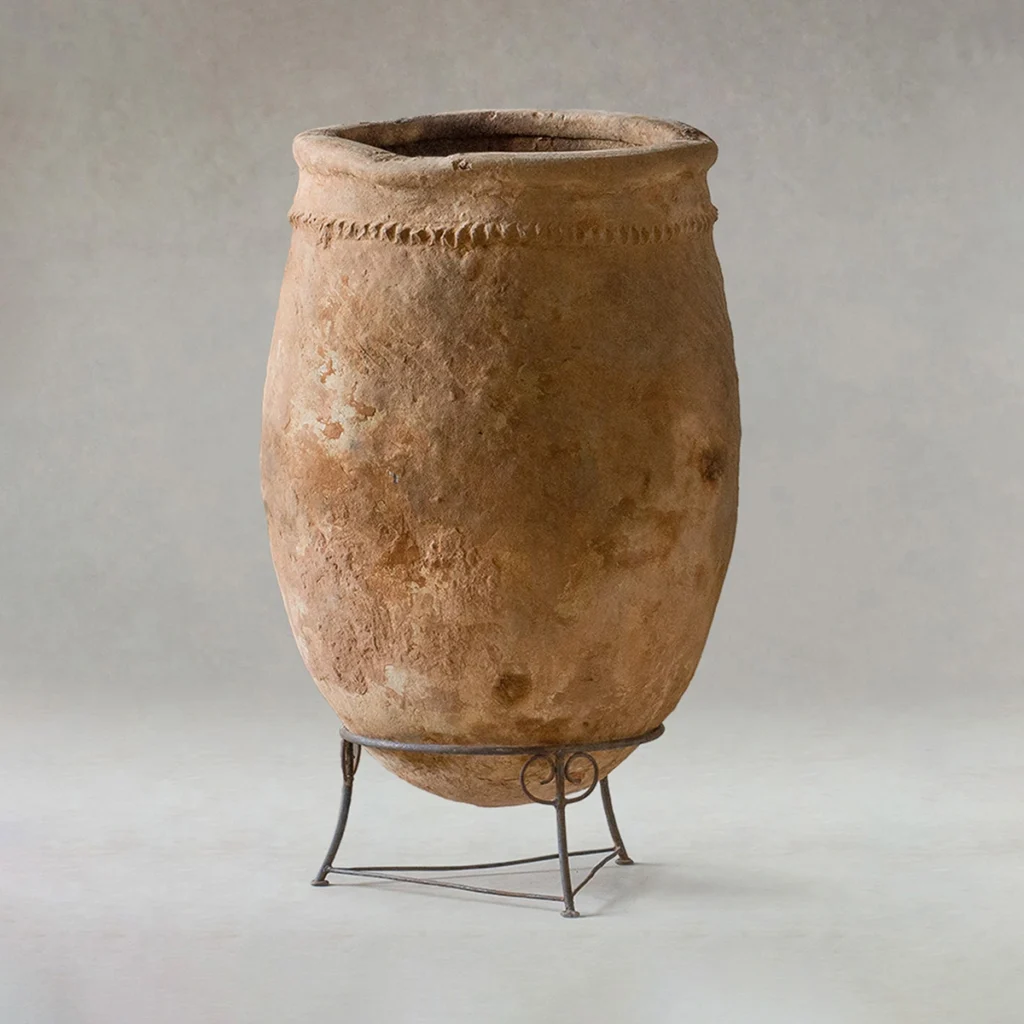 Large, weathered clay jar with a textured surface and simple rim, displayed on a metal stand against a plain background.