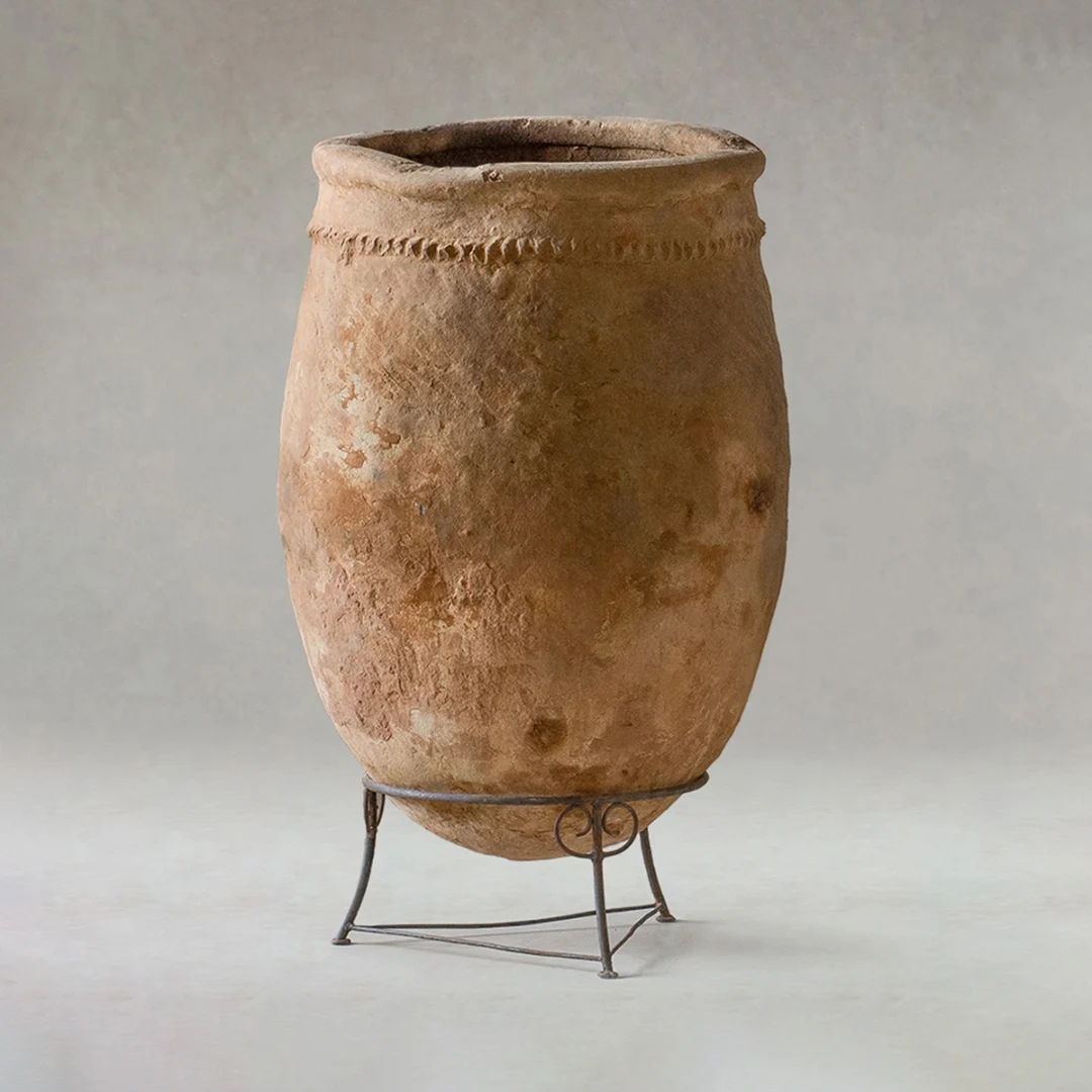 Large, weathered clay jar with a textured surface and simple rim, displayed on a metal stand against a plain background.