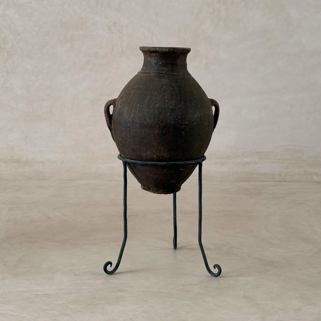 A dark, weathered clay pot with two handles is displayed on a simple black metal stand against a light, neutral background.
