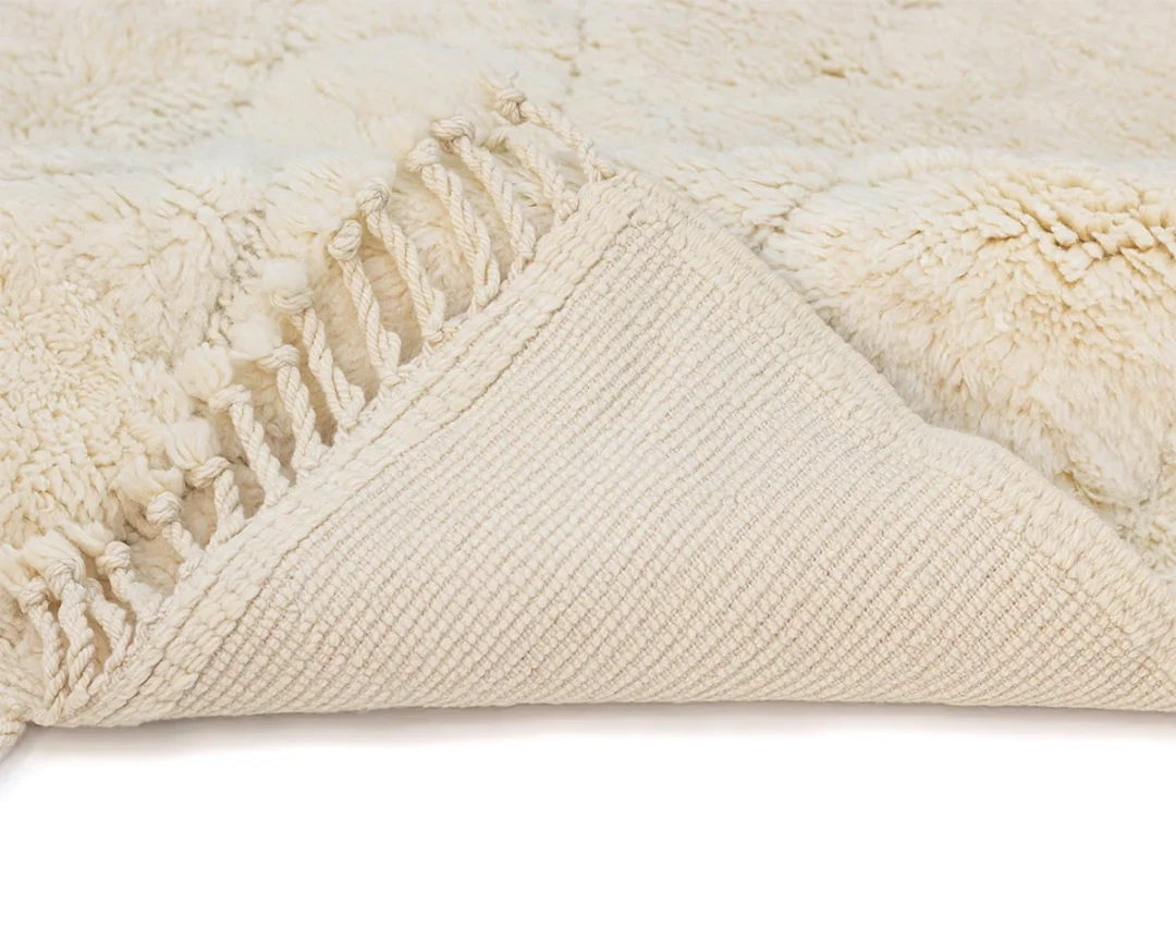 A close-up of a light-colored textured rug with fringed edges, partially folded to show the woven underside.