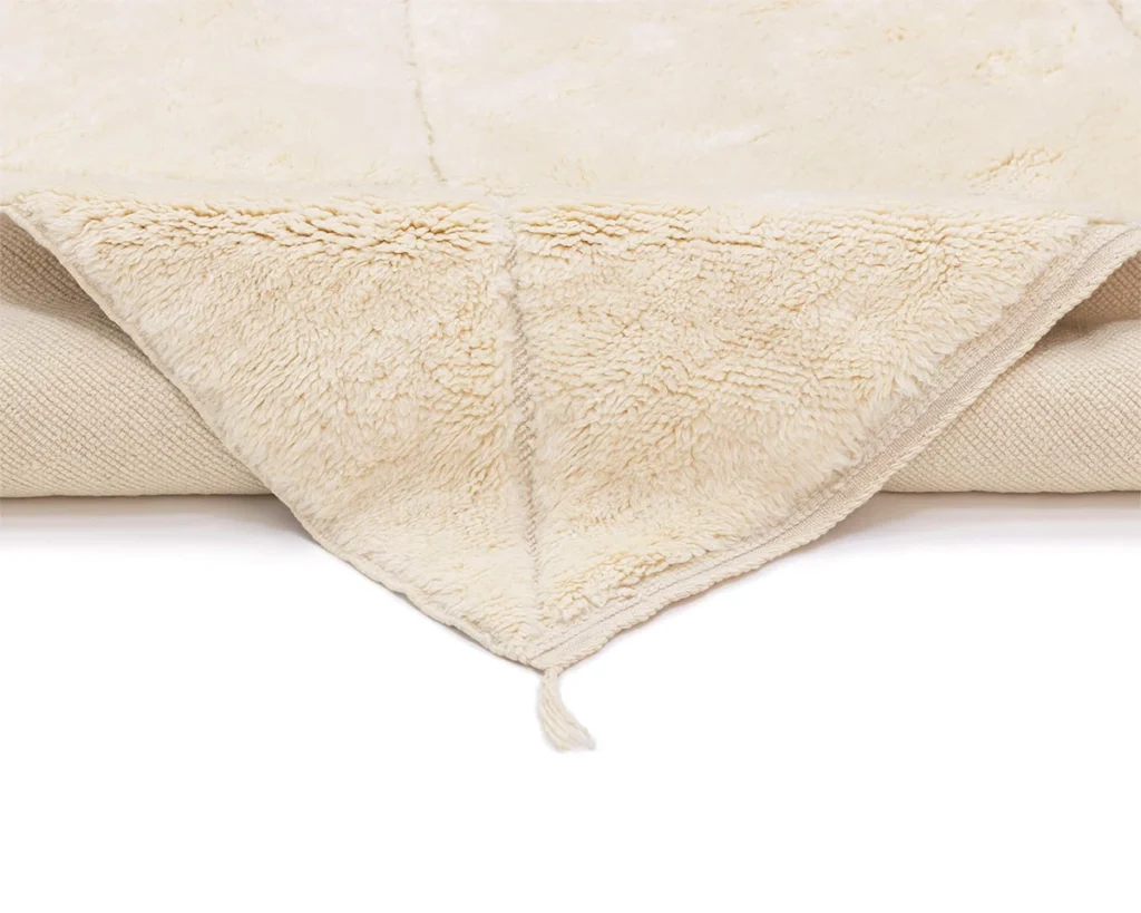 A close-up of a folded cream-colored fleece rug with a soft, fluffy texture and a small tassel at the corner, displayed on a white background.