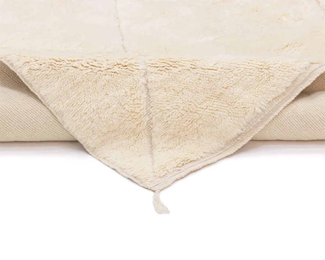 A close-up of a folded cream-colored fleece rug with a soft, fluffy texture and a small tassel at the corner, displayed on a white background.