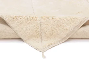 A close-up of a folded cream-colored fleece blanket with a soft, fluffy texture and a small tassel at the corner, displayed on a white background.