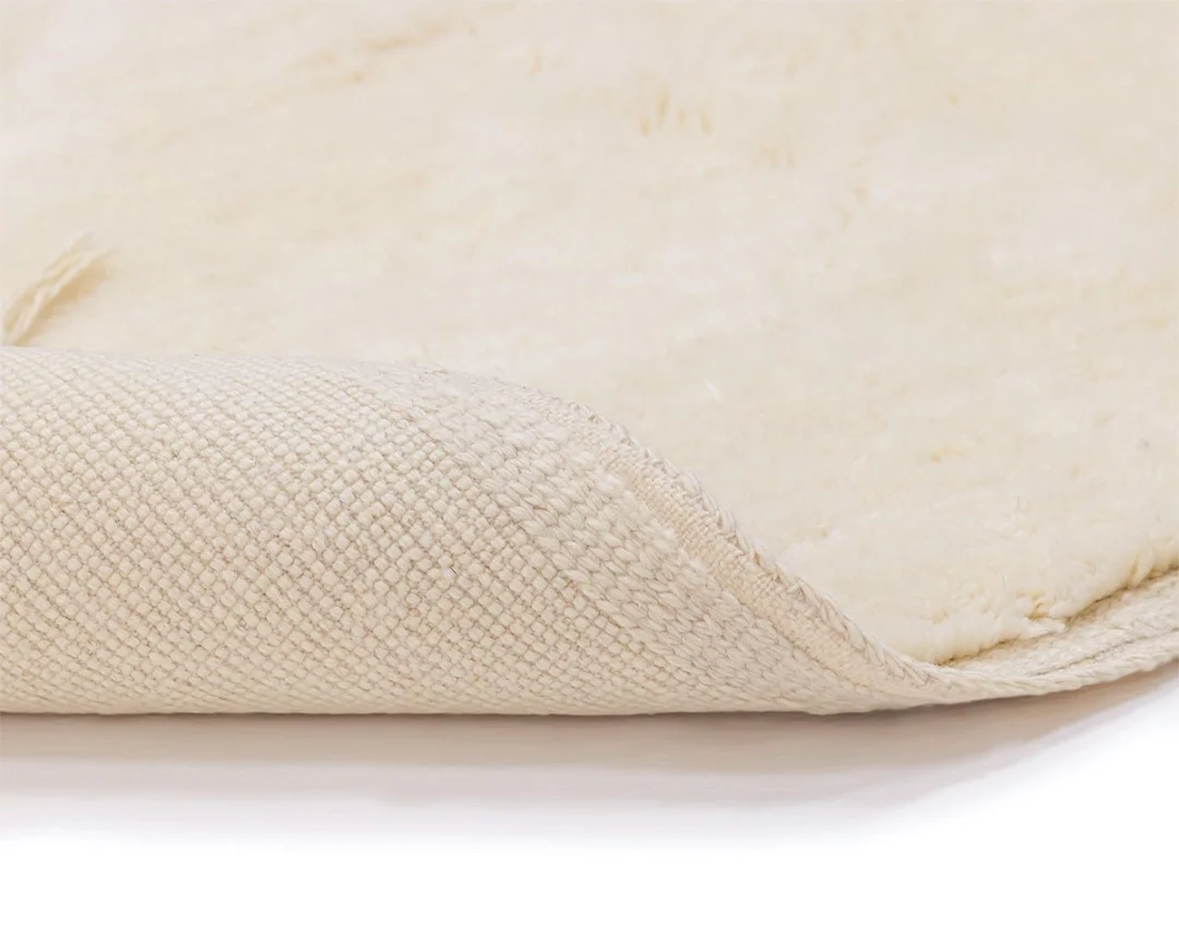 A close-up of a white or cream-colored area rug with the corner flipped over, showing the textured underside and soft, plush surface.