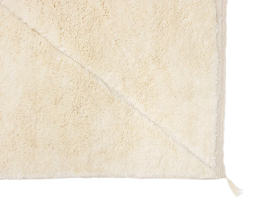 Close-up of a beige, fluffy area rug showing its soft texture and corner detail, with a small tassel at the edge.