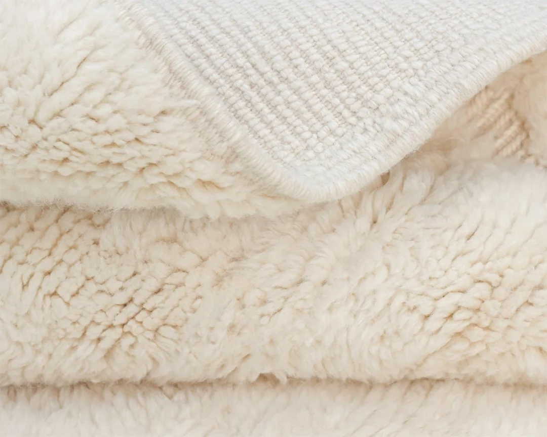 Close-up of a folded, cream-colored, fluffy fleece rug with a textured fabric edge visible on top.