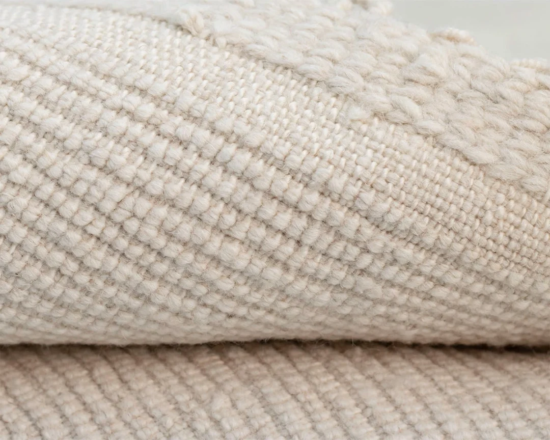 Close-up of a folded beige knotted rug with a textured, grid-like pattern and thick, raised yarn along the edge.