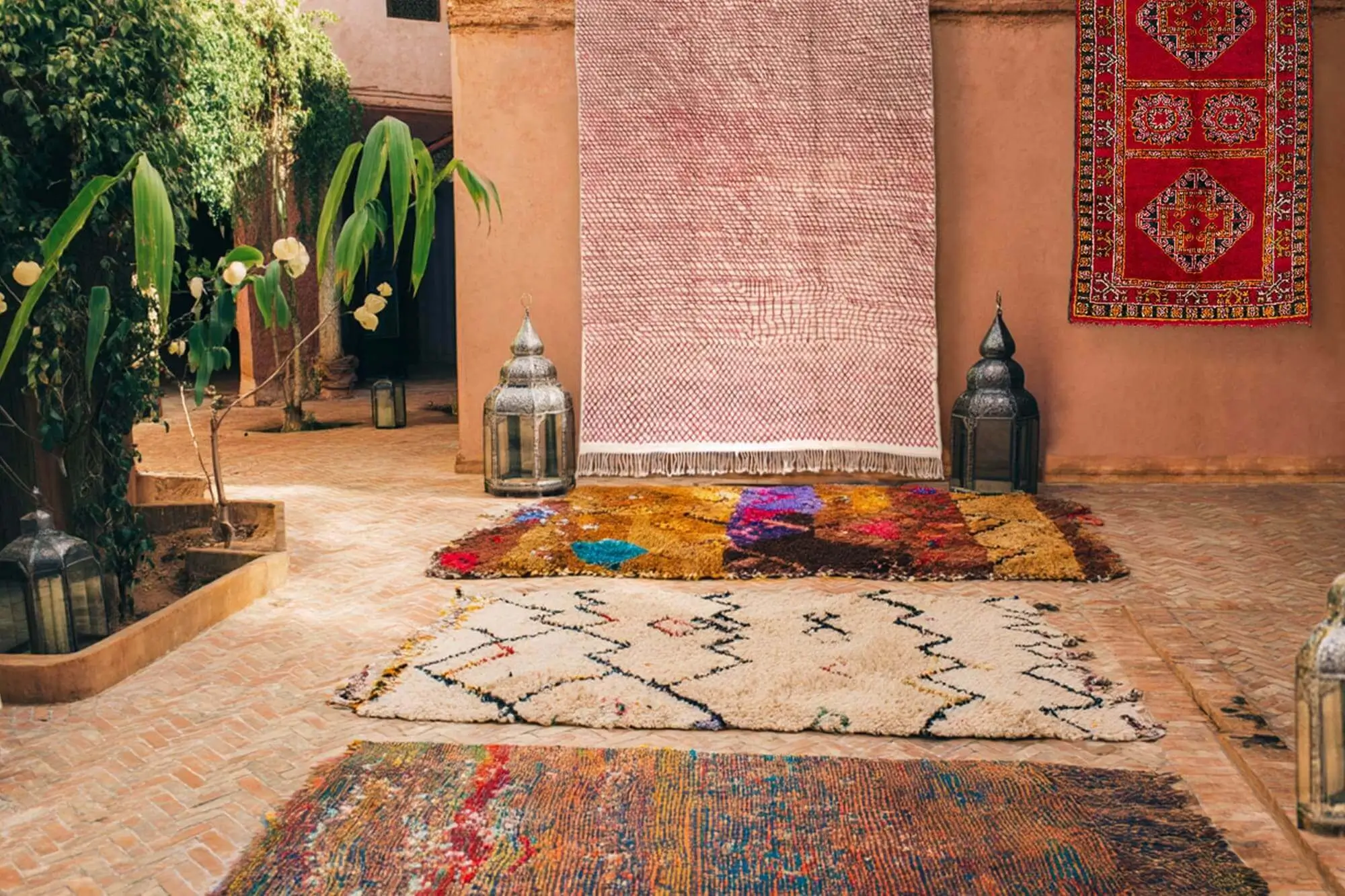 Three colorful rugs are laid on a brick patio near ornate lanterns and plants, with a pink woven textile hanging on a stucco wall and a red rug displayed to the right.