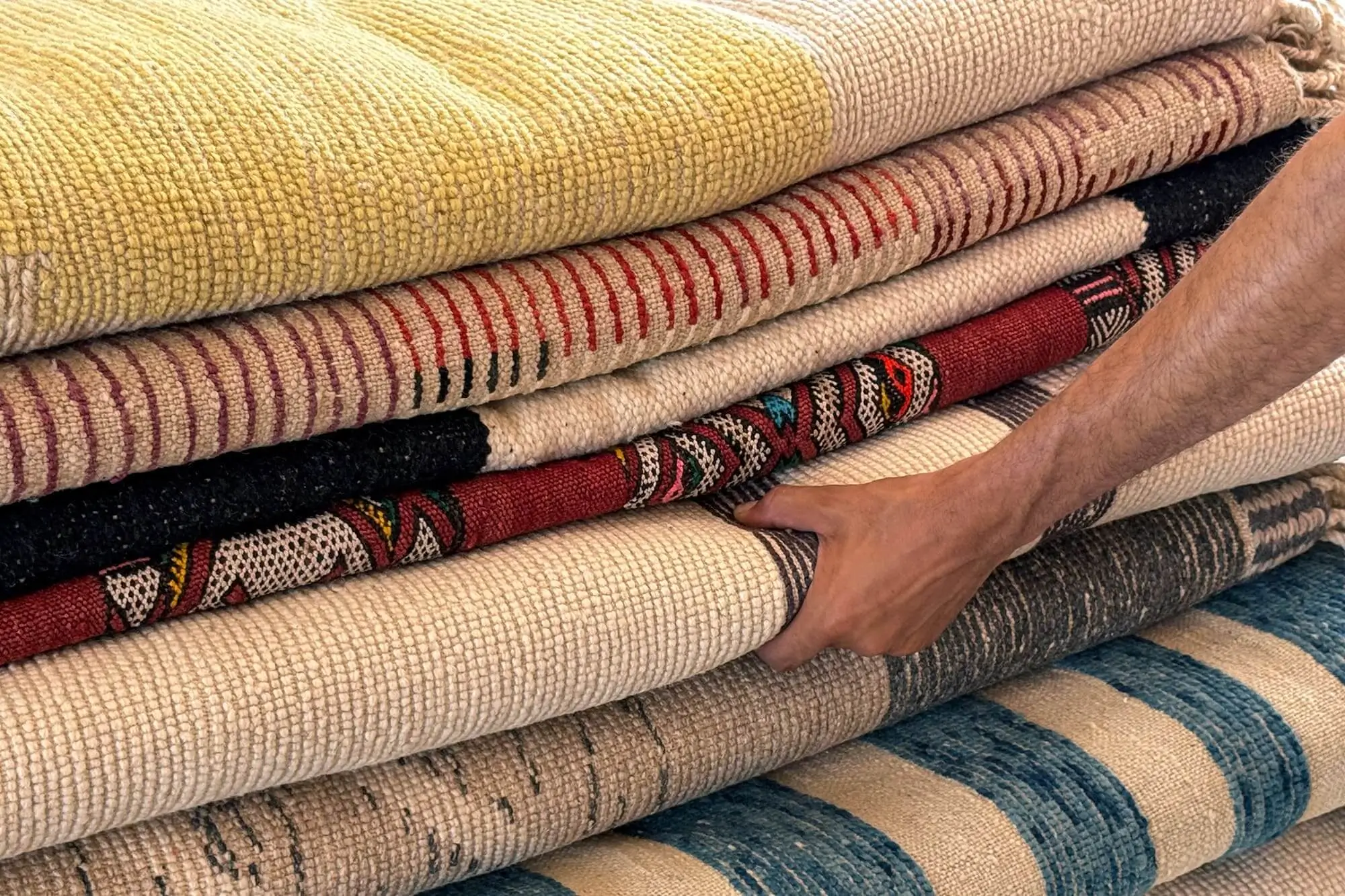A person’s hand lifts one of several stacked, patterned rugs in various colors and textures.
