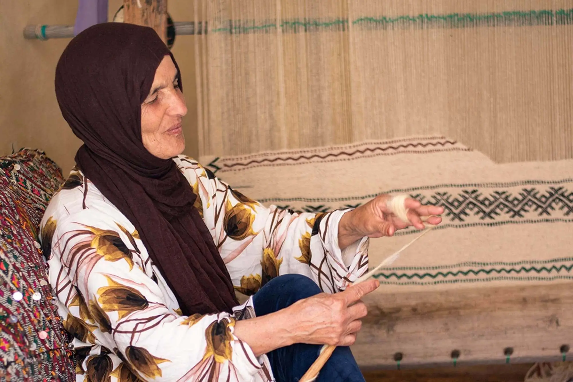 A woman wearing a floral-patterned shirt and a brown headscarf sits and spins thread by hand in front of a traditional loom.