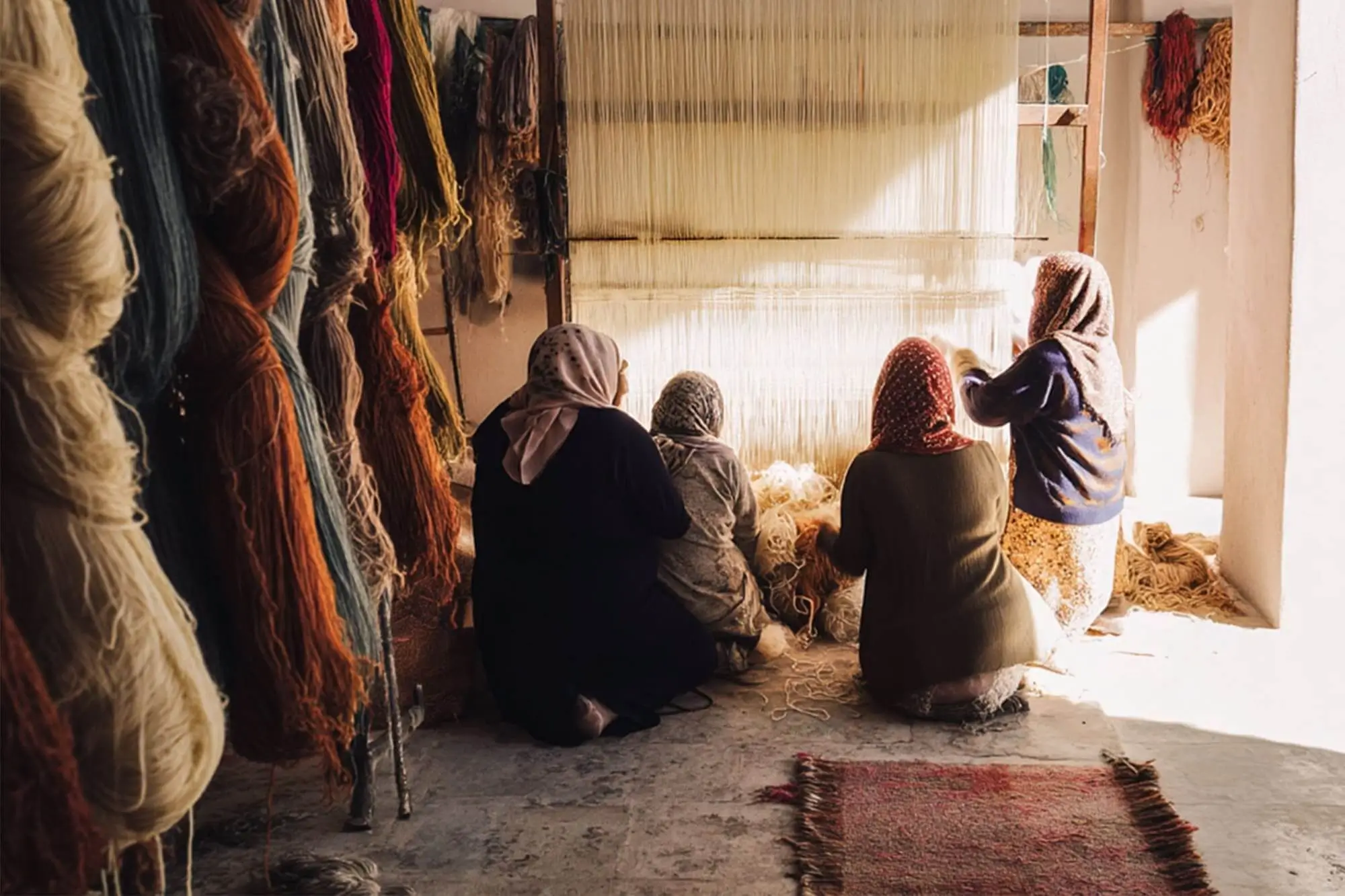 Four women sit on the floor, weaving at a large vertical loom in a sunlit room with bundles of yarn hanging on the walls.