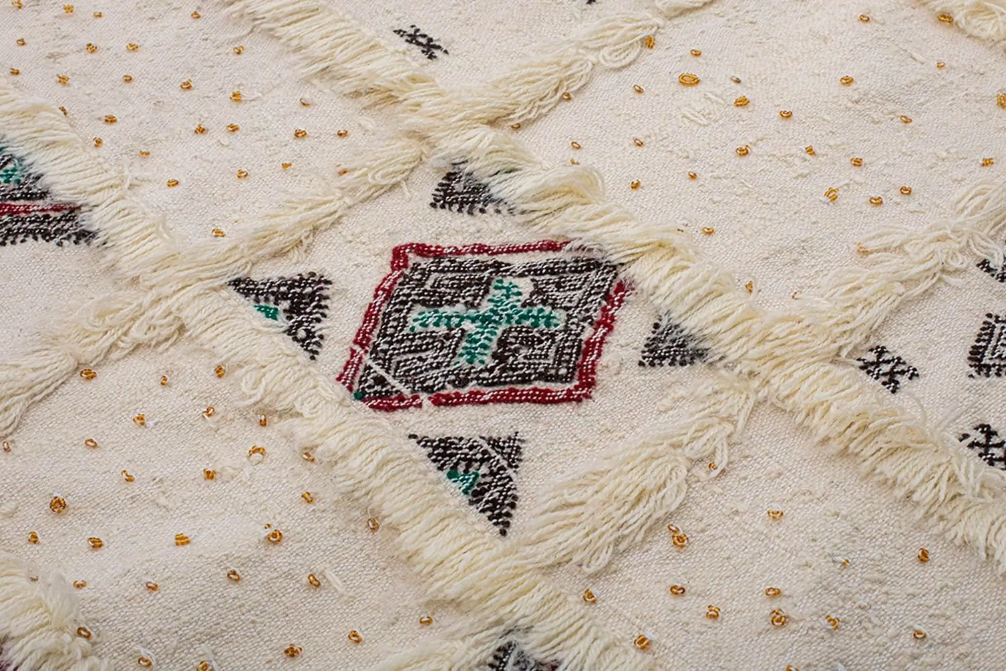 Close-up of a cream-colored textured fabric with diamond patterns, embroidered shapes in red, black, and green, and small gold decorative beads.