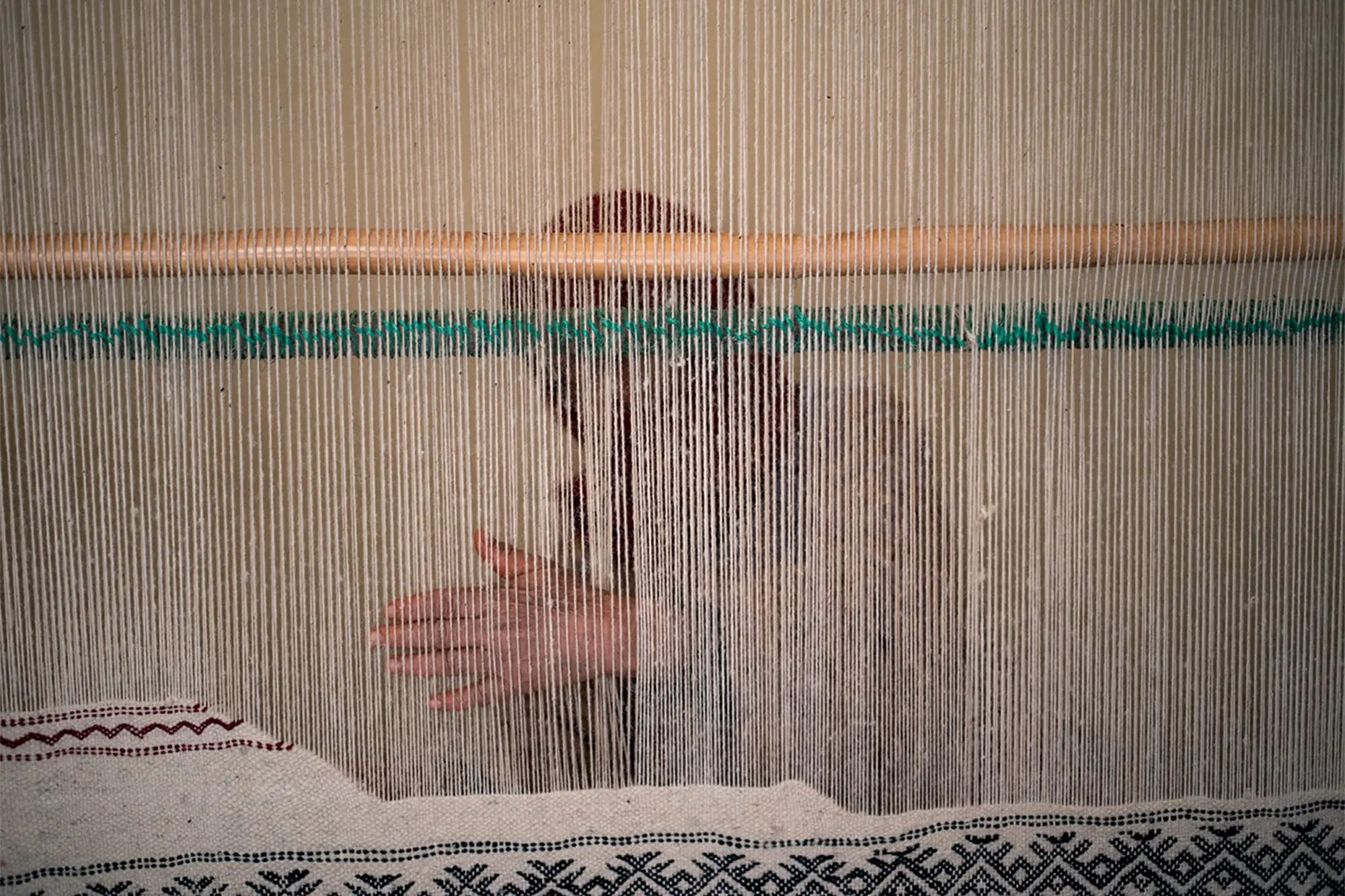 A women weaves fabric on a loom, partially obscured by vertical threads; a patterned textile is visible at the bottom of the loom.