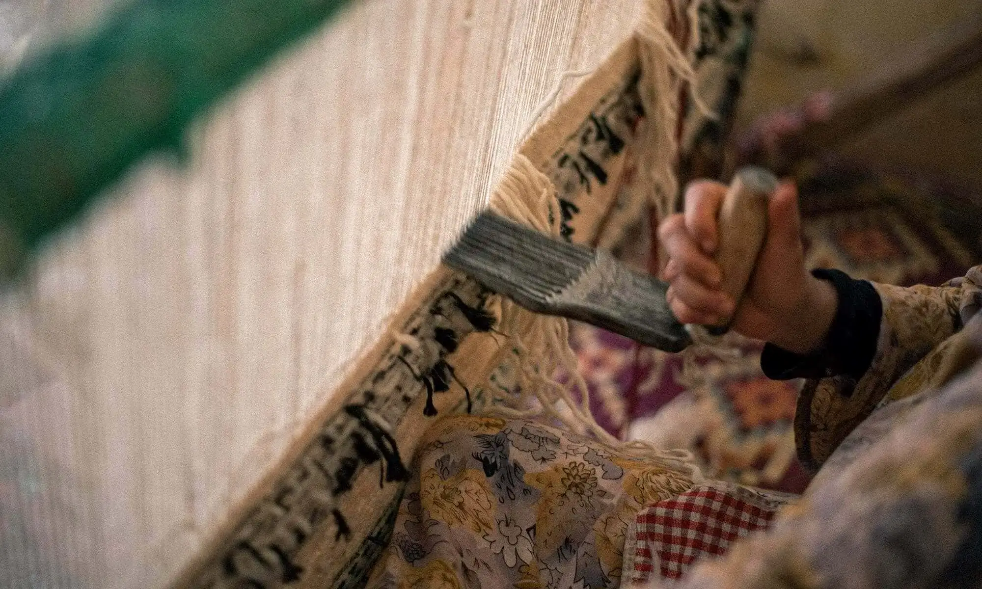 Close-up of a person weaving a carpet by hand, holding a tool against threads on a loom, with patterned fabric visible in the background.