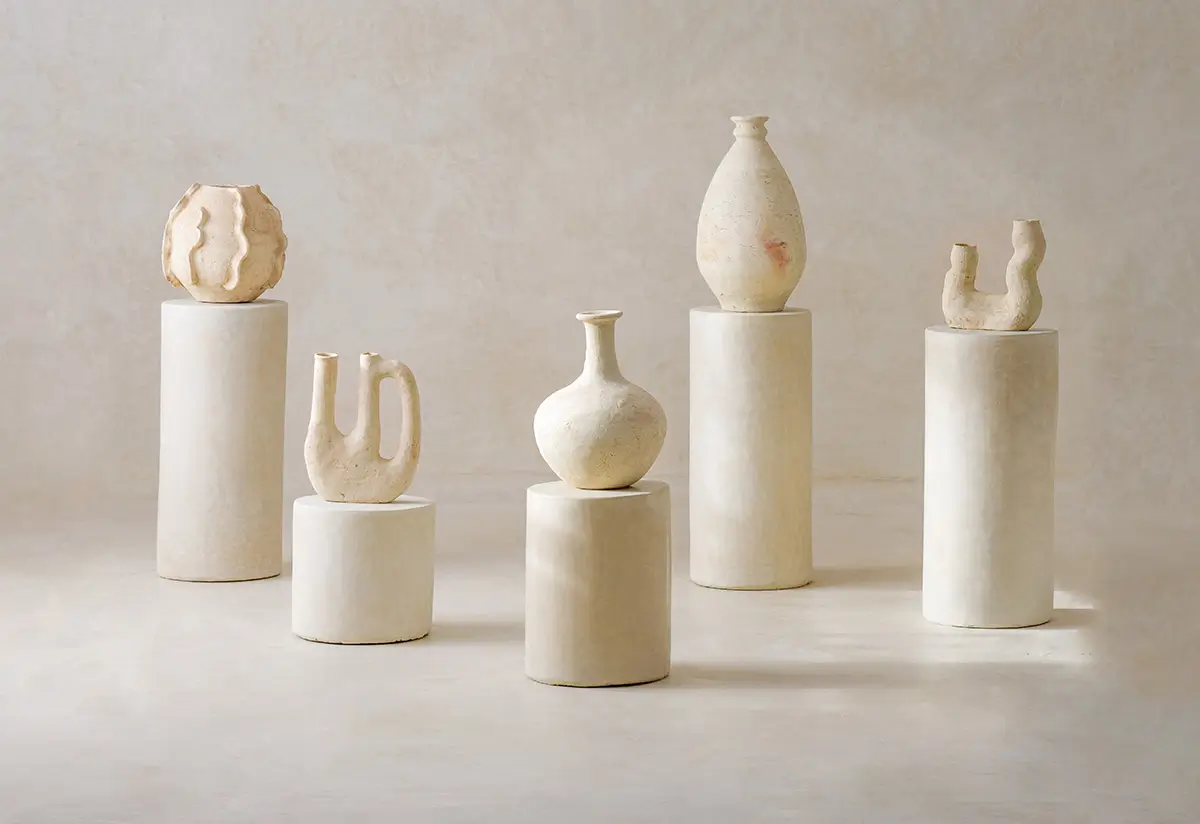 Six white or beige ceramic vases with varied shapes are displayed on cylindrical white pedestals against a neutral background, evoking the craftsmanship found in authentic Moroccan rugs.