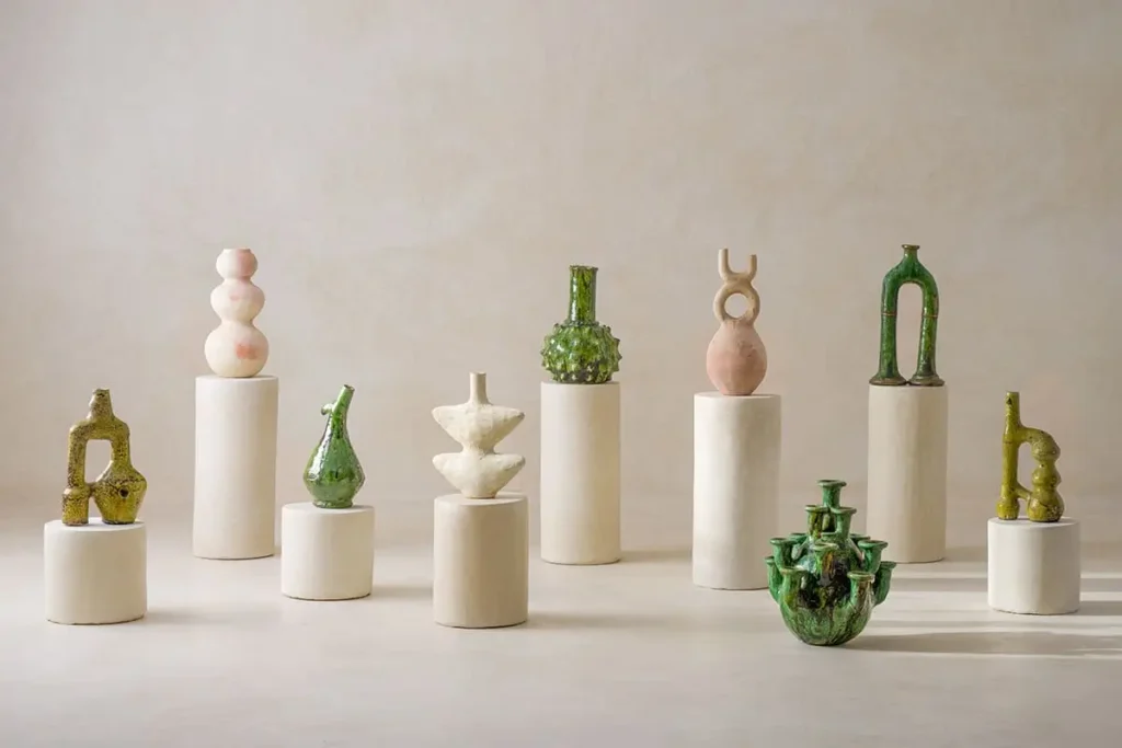 Ten uniquely shaped ceramic vases in neutral and green tones are displayed on white cylindrical pedestals against a plain beige background, creating an elegant scene reminiscent of spaces accented with authentic Moroccan rugs.