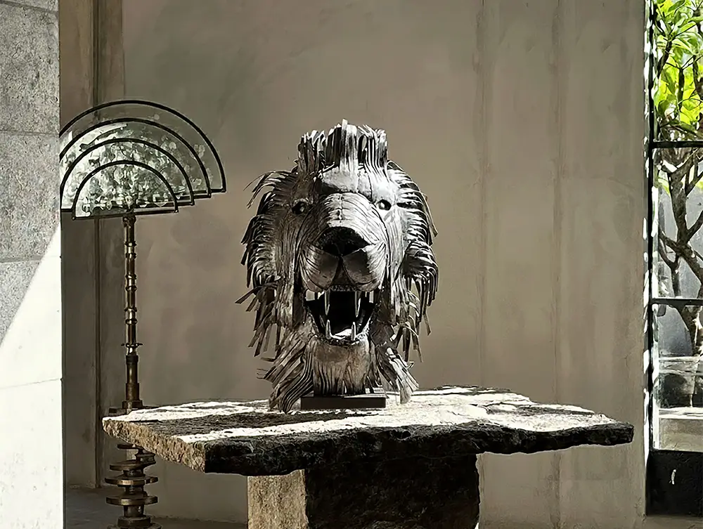 Azele Studio, Handmade Sculptures. A metal lion head sculpture with an open mouth sits on a stone table in a sunlit room featuring Authentic Moroccan Rugs by the large window. ethical home decor