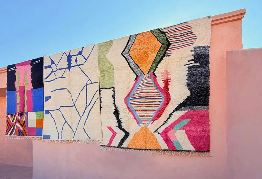 Three colorful, hand-knotted Authentic Moroccan Rugs with geometric patterns hang over a pink stucco wall under a clear blue sky.