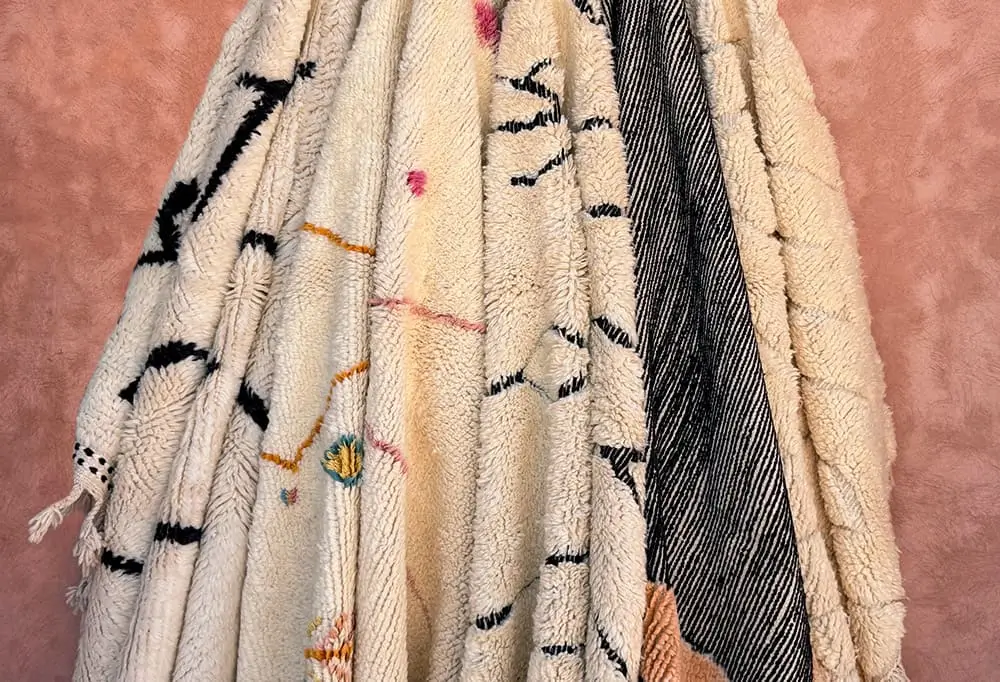 Several hand-knotted, authentic Moroccan rugs in neutral tones with black, orange, and pink accents hang side by side against a pinkish background.