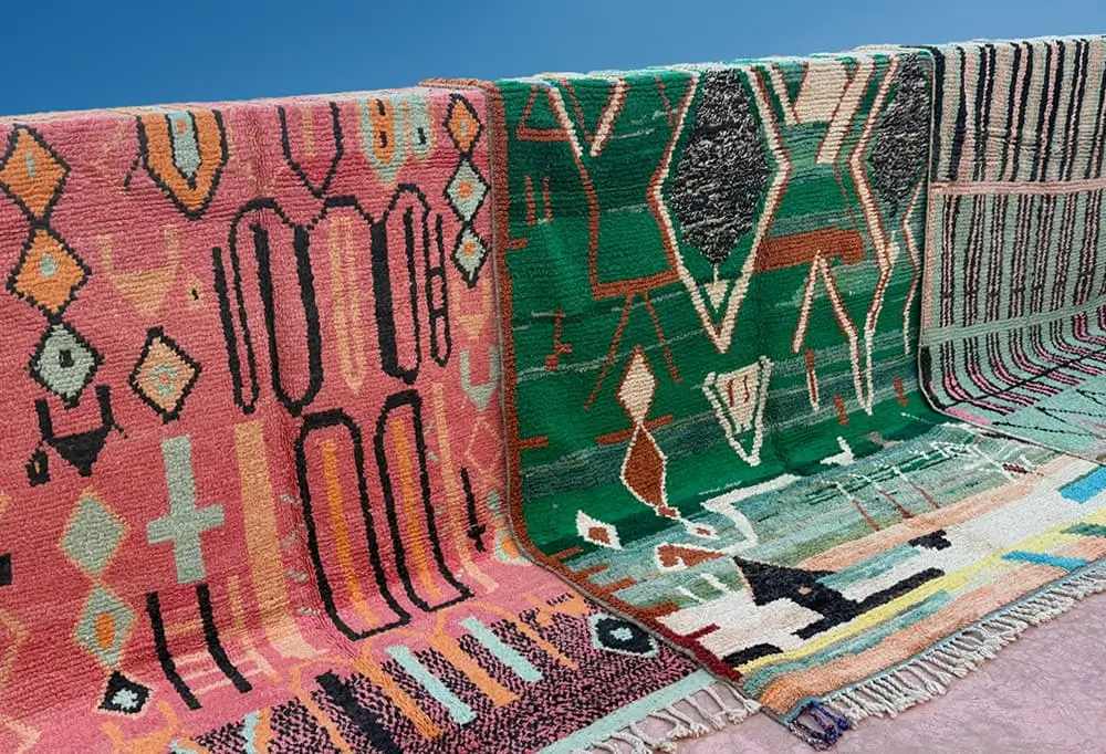 Three colorful, patterned hand-knotted rugs are draped side by side over a ledge against a blue background, showcasing the vibrant style of authentic Moroccan rugs.