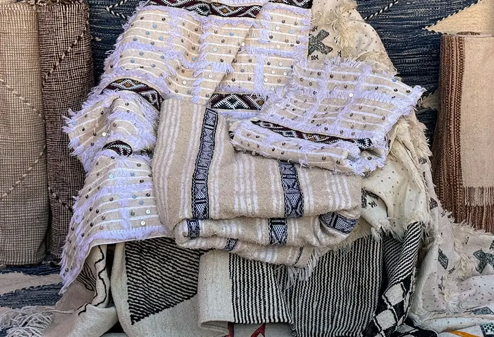 A collection of woven and textured rugs, including hand-knotted authentic Moroccan rugs, with various patterns and neutral colors, folded and stacked together.