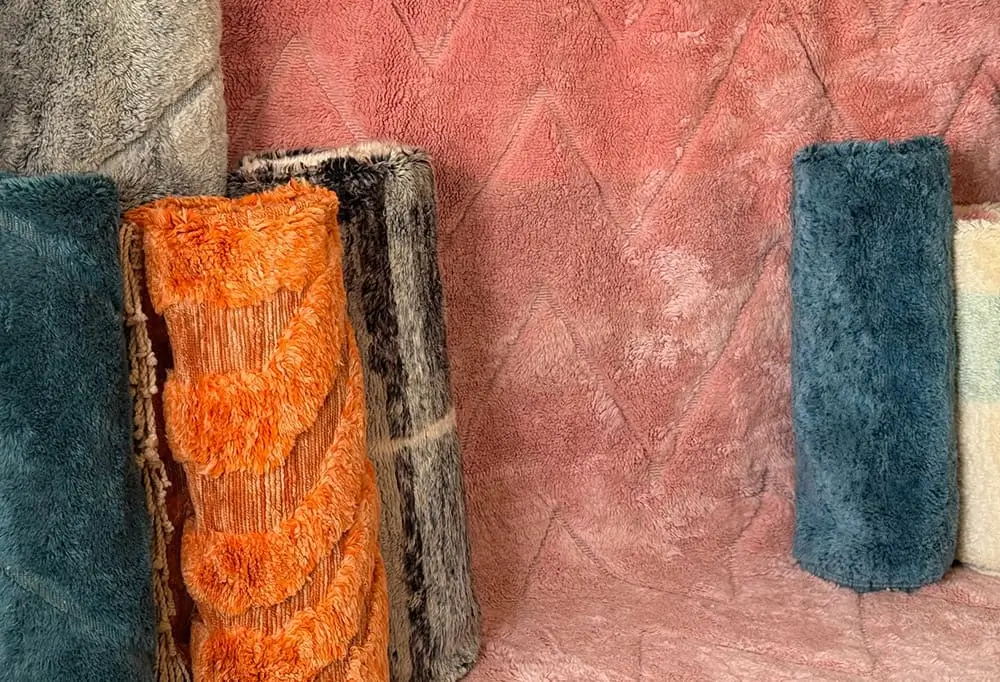 Five rolled-up shaggy rugs in various colors and patterns are displayed upright against a textured pink background.