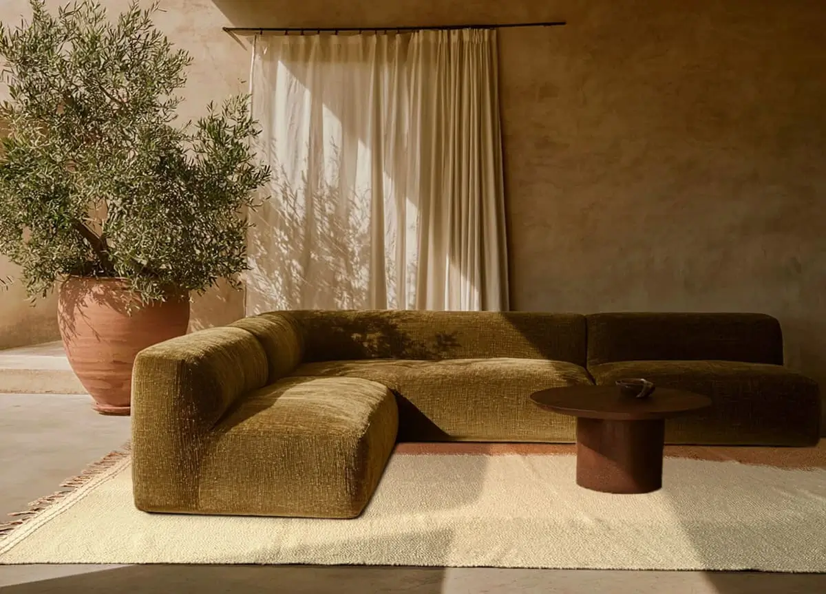 A green sectional sofa sits on a light rug beside a round wooden coffee table and a large potted tree, with cream curtains and a textured beige wall in the background, featuring an authentic Moroccan hand-knotted rug for added elegance.