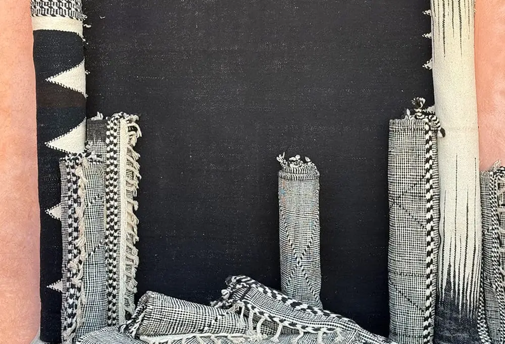 Several rolled and folded hand-knotted black and white patterned authentic Moroccan rugs are displayed upright against a plain black background.