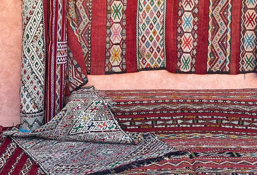 Authentic Moroccan rugs with colorful, hand-knotted geometric patterns are displayed as vibrant wall hangings against a pink wall.