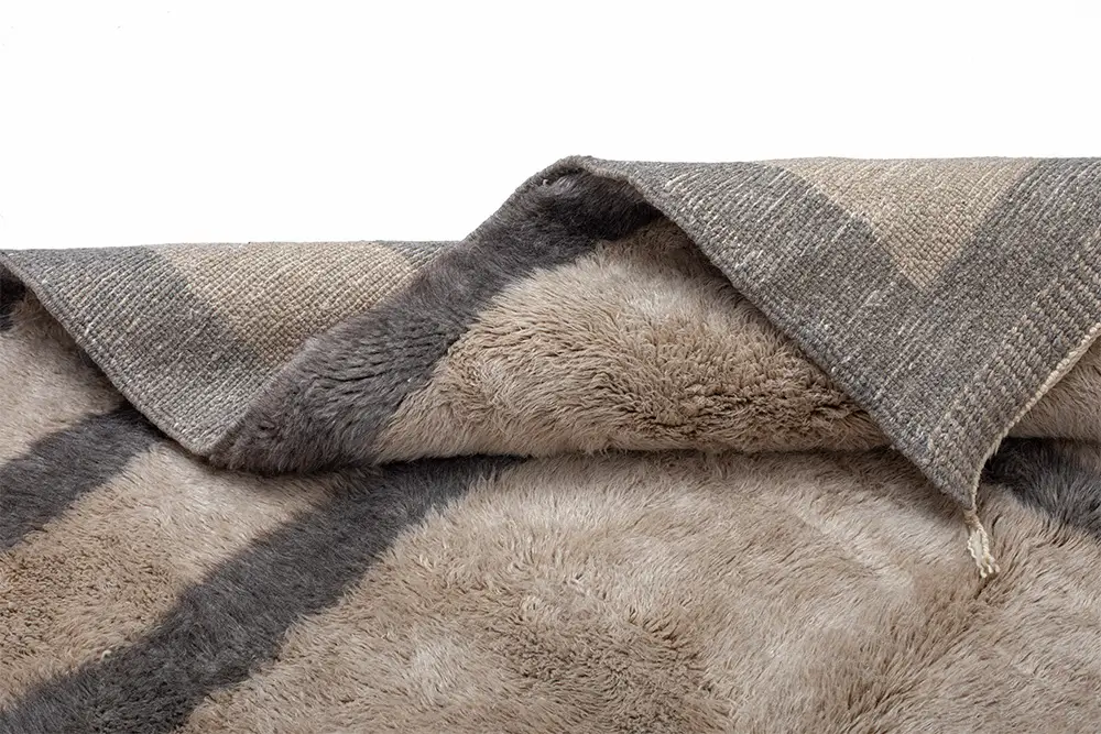 Discover the charm of this authentic Moroccan rug's detail, featuring taupe and gray stripes with a textured, fluffy surface. A folded corner reveals its artisanal quality, showcasing both beauty and craftsmanship. Perfect for adding warmth to any room.