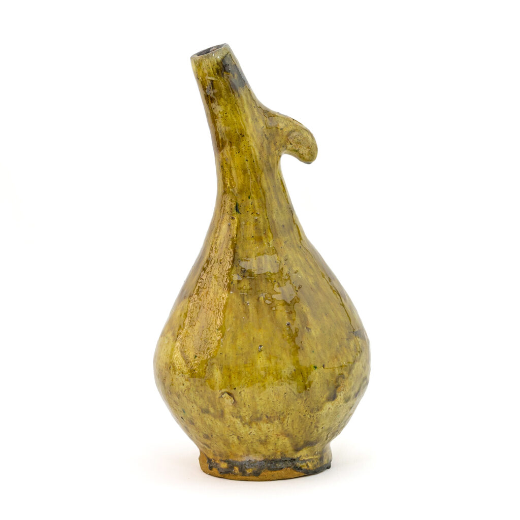 An ancient, beige-yellow Tamegroute pottery vessel with a curved spout and handle, featuring a rounded base and a weathered surface.