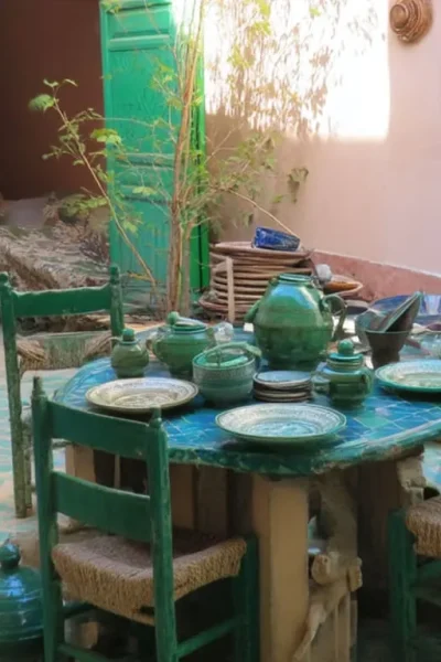 Table with green pottery set in a courtyard with patterned floor, surrounded by green chairs. Assorted pottery and woven items are visible on the floor and walls.
