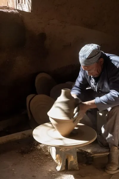 A person in a dimly lit workshop shapes a clay pot on a potters wheel surrounded by stacks of pottery. Sunlight filters through a window, illuminating the scene.